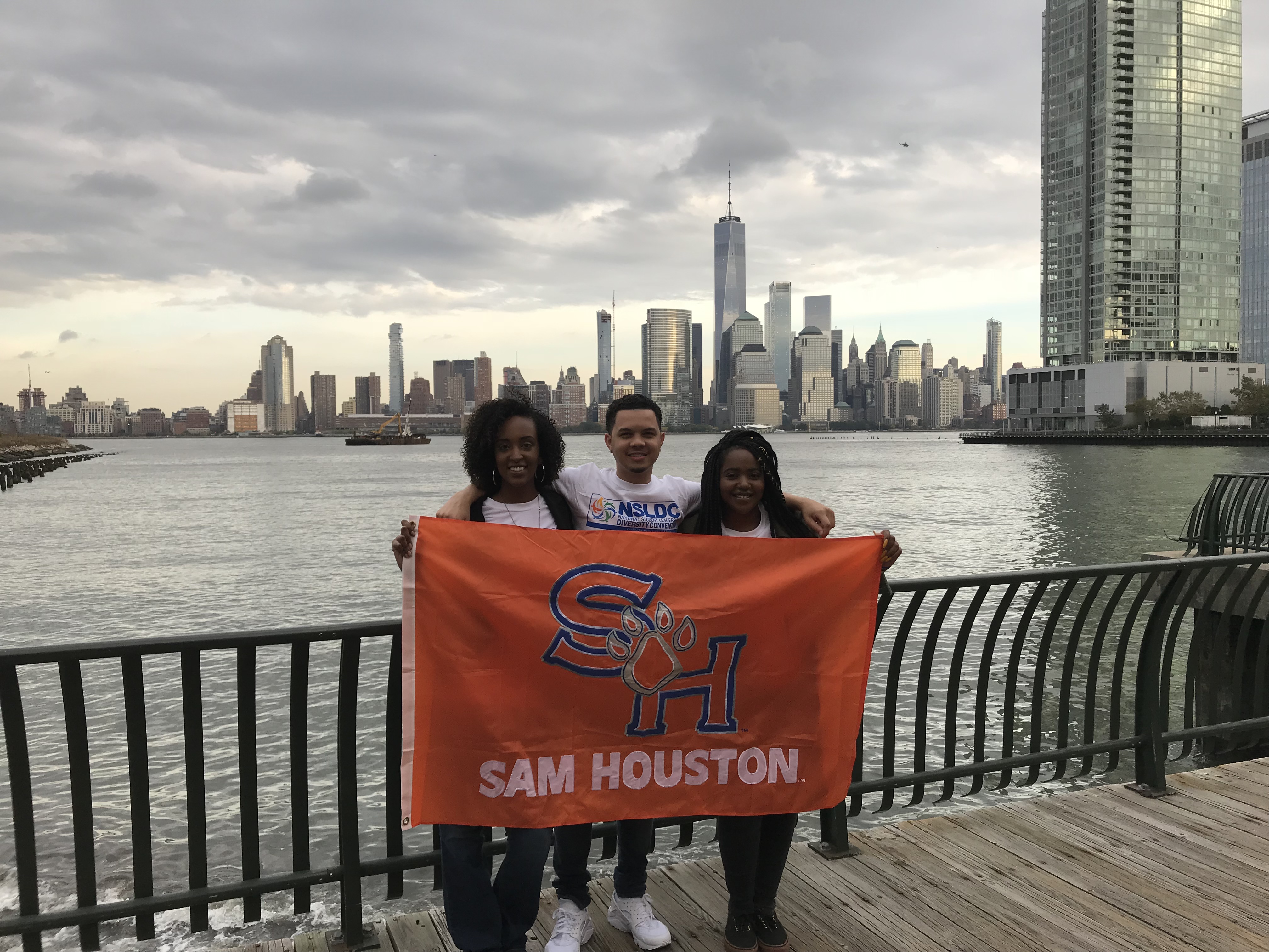 Students pose on a bridge holding a SHSU flag. There is a large body of water behind them and the One World Trade center in New York City is in the skyline in the distance.
